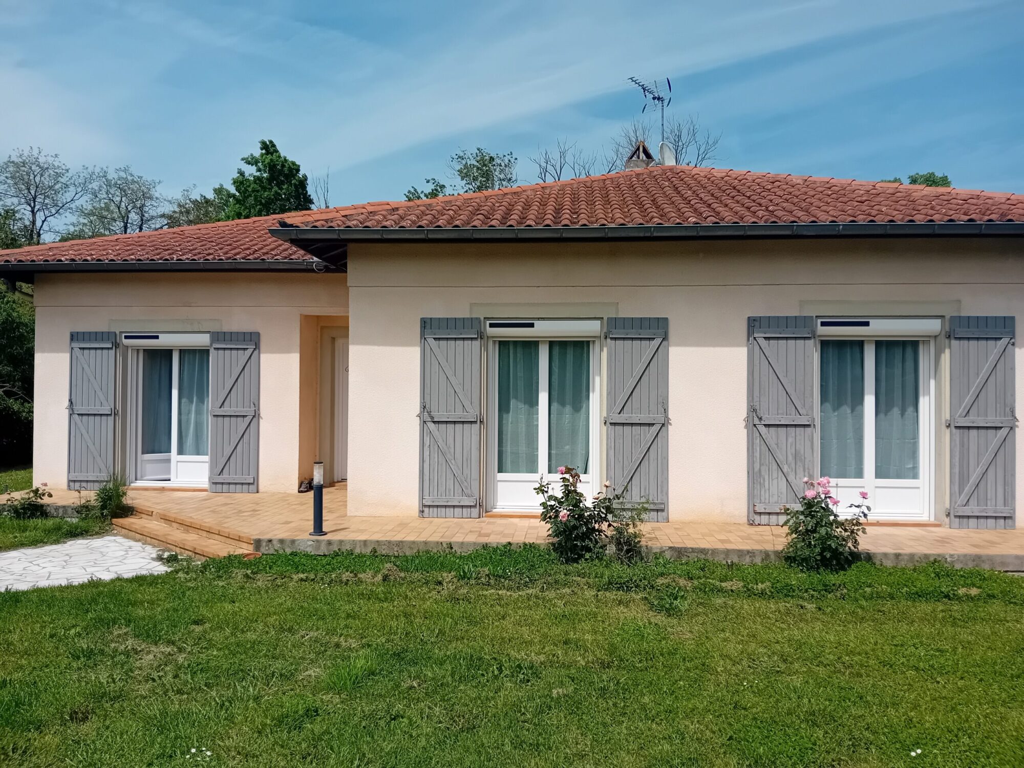 Façade de maison avec volets en menuiserie grise et fleurs au jardin.